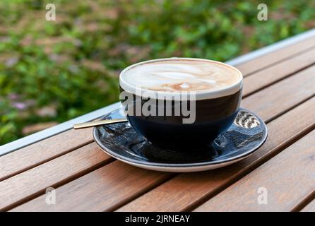 cappuccino o caffè latte in tazza nera sul tavolo di legno all'esterno Foto Stock