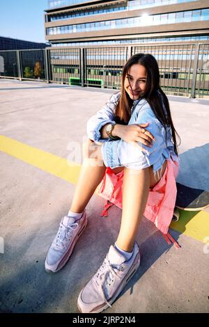 Una ragazza skater adolescente siede alla stazione ferroviaria sullo skateboard e sorride alla macchina fotografica. Foto Stock