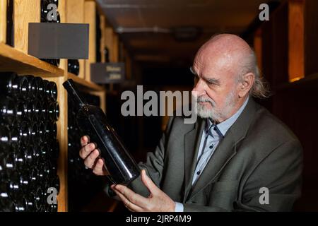 Senior man in cantina con bottiglie in background bere e degustare vino. Foto Stock