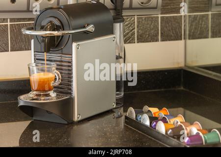 Macchina per caffè a capsule su un piano di appoggio nero, e una tazza trasparente che riempie di caffè e capsule di vario tipo Foto Stock