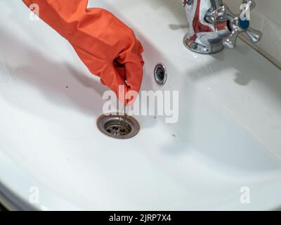 La mano nei guanti arancioni tiene molte perdite di capelli sul filtro nel lavabo durante la pulizia Foto Stock