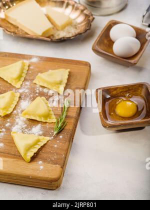 Ravioli grezzi triangolari fatti in casa su un tagliere con rametto di rosmarino, ingredienti, utensili da cucina. Isolato su sfondo bianco. Cucina italiana. Reg Foto Stock