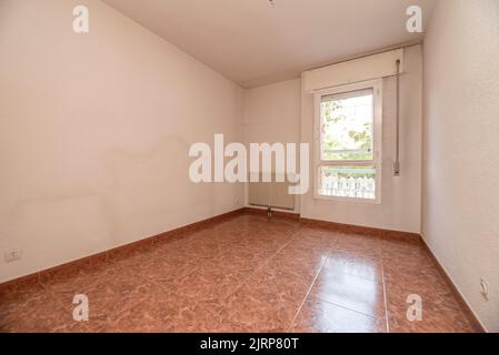 Soggiorno vuoto con pavimento in gres rossastro, radiatore bianco a parete e finestra con vista Foto Stock