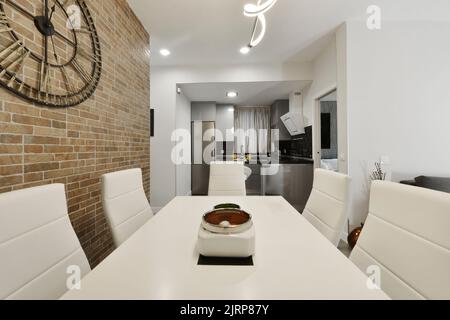 soggiorno con parete in mattoni a vista, tavolo da pranzo in legno bianco con sedie tappezzate in similpelle e cucina aperta sullo sfondo Foto Stock