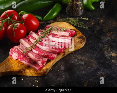 Bistecche di carne cruda con un rametto di rosmarino su un tagliere di legno, verdure su sfondo scuro. Cucinare bistecche di manzo, maiale nel cucinino casa Foto Stock