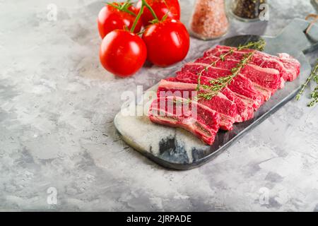 Su sfondo chiaro, affettate bistecche di carne cruda su un tagliere con un rametto di rosmarino, pomodori maturi, spezie, condimenti in vasetti di vetro. Ricette per Foto Stock