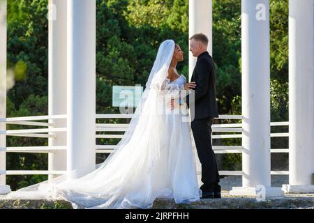 Sposi novelli a razza mista in una passeggiata che abbraccia lo sfondo di un bellissimo gazebo Foto Stock