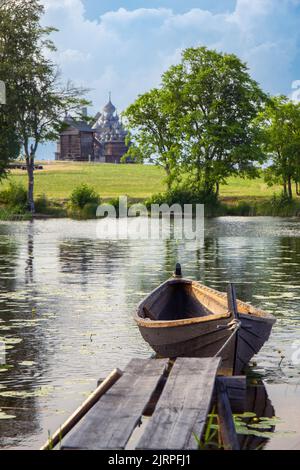 Paesaggio sull'isola di Kizhi nel lago Onega in una giornata di sole estivo. Vecchia barca di legno da pesca Kizhanka sulla riva. Villaggio con mulino d'epoca, tempio e chiesa Foto Stock