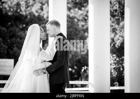 Sposi novelli a razza mista in una passeggiata che abbraccia e guarda amorevolmente l'un l'altro, fotografia in bianco e nero Foto Stock