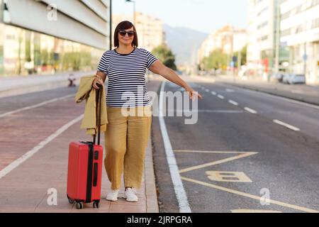 Donna d'affari anziana sorridente in occhiali da sole con valigia rossa che solleva la mano per prendere un taxi, taxi al terminal o alla stazione della città moderna. Viaggi bu Foto Stock