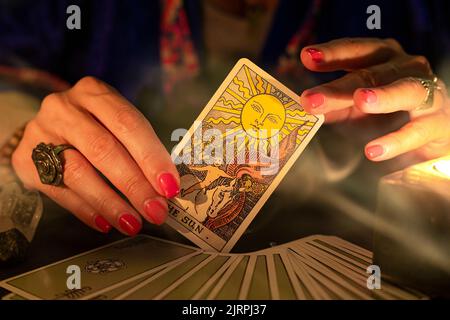 Fortune Teller mani femminili che mostrano la carta del tarocco del Sole, simbolo di positività e ottimismo, durante una lettura. Primo piano con luce a candela e fumo, moo Foto Stock