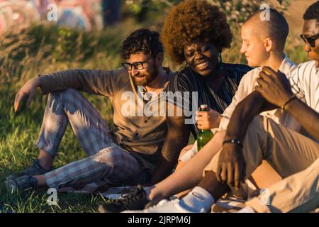 vicino mentre si siede sull'erba, bevendo birra, parlando e ridendo. Vacanze estive. Foto di alta qualità Foto Stock
