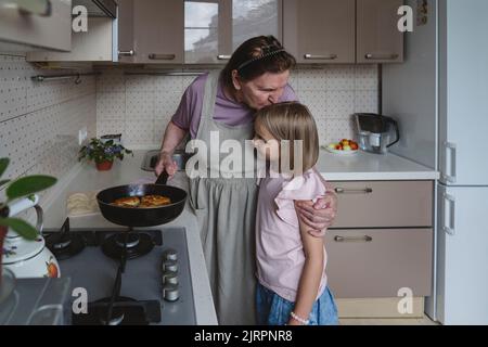 Una donna anziana in cucina frigge torte con la nipote. Foto Stock