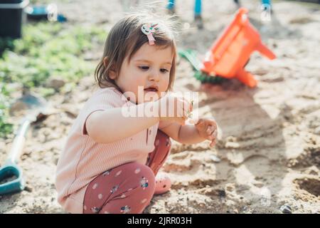 Una bambina caucasica piccola in piedi nel giardino tenendo una pianta nelle sue mani e la esamina. Natura estiva. Bella ragazza giovane. Giorno di sole. Foto Stock