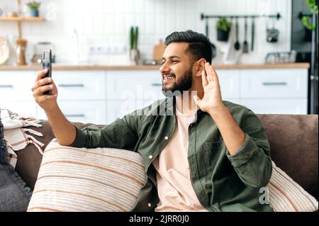 Positivo misto razza giovane uomo elegante seduto sul divano in salotto, utilizzando il suo smartphone, parlando in videochiamata. Buon video blogger indiano o arabo, fa un selfie telefono, registra un flusso, sorriso Foto Stock