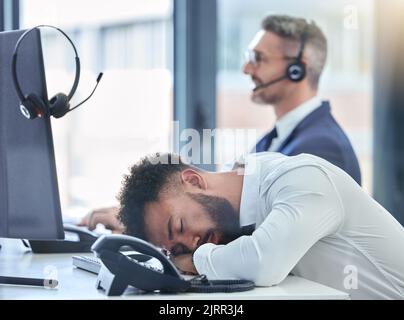 Un operatore del call center pigro, addormentato e stanco al suo tavolo o alla sua scrivania al lavoro ha lavorato soffrendo di burnout. Impiegato giovane del servizio clienti esaurito Foto Stock