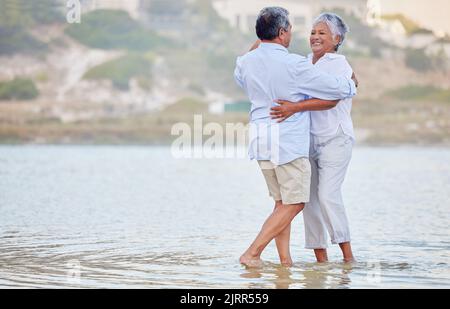 Paesaggio, spiaggia e danza di una coppia anziana sorridente e danzante in mare o acqua dell'oceano e abbracciante al tramonto. Felice, sorriso o legame di un vecchio Foto Stock
