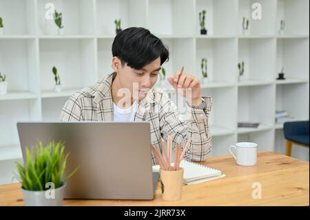 Concentrato giovane asiatico studente universitario maschile o freelance lavorando e progettando il suo progetto nella sua stanza di lavoro. Foto Stock
