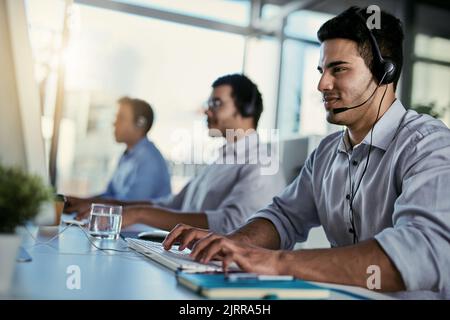 Su chiamata e pronto a fornire un'assistenza clienti efficiente. Un operatore del call center che lavora in ufficio con i colleghi in background. Foto Stock