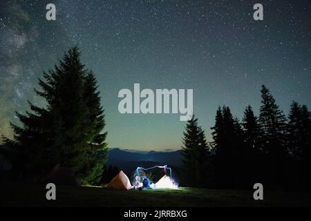 Luminose tende turistiche illuminate che brillano sul campeggio in montagne scure sotto il cielo notturno con stelle scintillanti. Concetto di stile di vita attivo Foto Stock