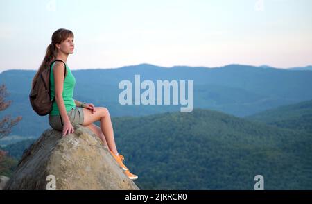 Sportiva donna escursionista seduto da solo facendo una pausa sul sentiero collinare. Donna solitaria che gode di vista della natura serale dalla scogliera rocciosa sul sentiero selvaggio Foto Stock