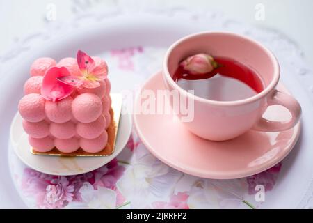 Serve un vassoio festoso di appetitosa torta cremosa con decorazione a farfalla e tazza rosa di fragrante tè gustoso con petali di fiori primo piano. Rosa Foto Stock