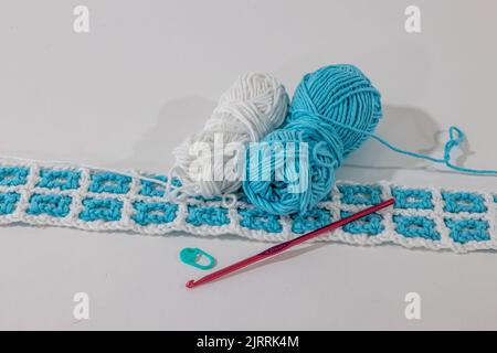 Tessuto intrecciato a crochet nei colori bianco e blu accanto a due palle di cotone, gancio a crochet rosa metallizzato e pennarello a punti su sfondo bianco, Foto Stock
