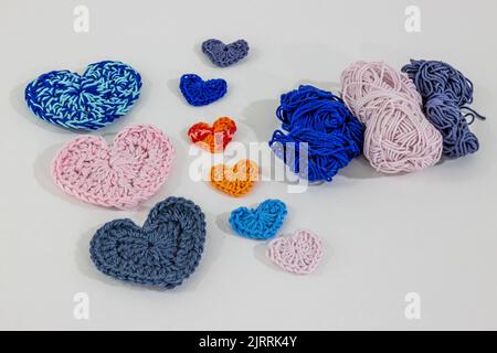 Cuori di crochet di diversi colori e dimensioni accanto a palle di cotone blu, rosa e grigio su una superficie e sfondo bianco, anche in arancio e misto colo Foto Stock