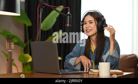 Donna che indossa una cuffia che parla in un microfono mentre crea un podcast online dal suo studio di casa Foto Stock