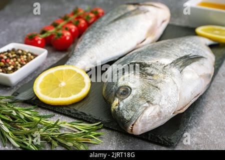 Pesce crudo dorado su tagliere di ardesia nera e fondo in cemento grigio con spezie, pomodoro, rosmarino, olio d'oliva e limone Foto Stock