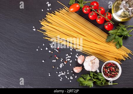 Pasta e ingridienti di cottura su superficie di ardesia nera. Spaghetti, pomodoro, olio d'oliva, spezie, aglio e prezzemolo. Concetto di cucina italiana. Vista dall'alto, piatta Foto Stock