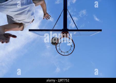 L'uomo sta allenando il basket sul campo di strada. Lancia la palla al cerchio. Angolo basso Foto Stock