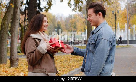 Ragazzo caucasico dare scatola rossa piccolo regalo con nastro d'oro a bella ragazza giovane donna in autunno parco sorpresa presente per San Valentino Foto Stock