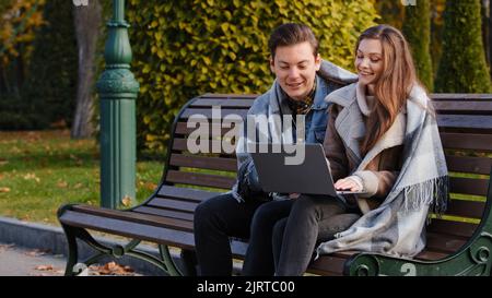 Coppia freelance due partner gente giovane donna caucasica e uomo imprenditori sedersi su panca in parco uso urbano wifi lavoro sul laptop guardare dispositivo Foto Stock