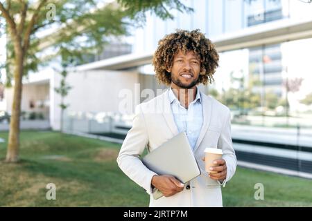 Ritratto di positivo bel brasiliano o ispanico capelli ricci uomo, ceo, top manager, agente immobiliare, tenendo notebook e bicchiere di caffè, stand fuori vicino al business center, guarda la fotocamera, sorriso Foto Stock