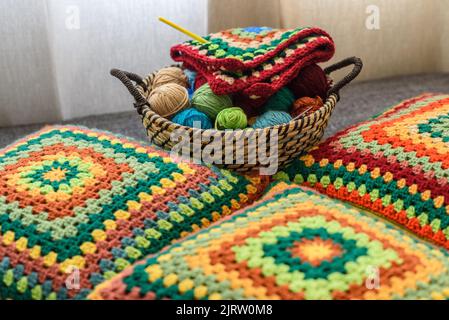 Uncinetto quadrato Granny. Creazioni fatte in casa. Filati di lana colorati in un cesto di paglia su un divano. Lavorazione a maglia fatta a mano, cuscini in maglia e un'atmosfera carina. Foto Stock