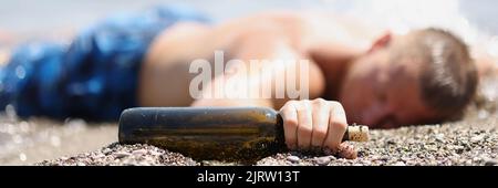 Il giovane è sdraiato sulla spiaggia con una bottiglia di alcool Foto Stock