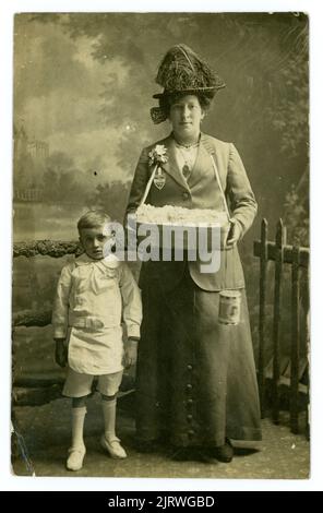 Cartolina originale, chiara, ritratto in studio di una donna anziana, matura, collezionista volontario di beneficenza. È una venditrice di rose di beneficenza per l'Alexandra Day e il bambino accanto a lei è probabilmente un bambino povero o orfano di Bethnal Green. Il bambino è vestito in modo intelligente ma con un vestito bianco improprio e scarpe troppo piccole. La donna tiene in mano una scatola da collezione che contiene il patrono, il ritratto della regina Alessandra. La donna indossa un distintivo che indica di essere una collezionista ufficiale. Dallo studio di A. Lewis 228 Green St. Bethnal Green, Londra, U. K circa 1913. Foto Stock