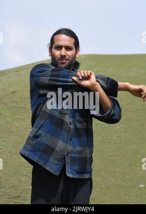Vista frontale di un giovane ragazzo che fa Cross Body Shoulder Stretch esercizio all'aperto. Un ragazzo indiano caldo con spalla croce braccio stretch posa. Foto Stock