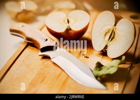 Su una tavola da cucina di legno si trova una mela matura intera e mezza mela, e accanto ad essa un coltello affilato e foglie illuminate dalla luce del sole. Nutritio sano Foto Stock