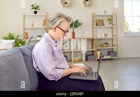 Donna anziana di successo con i capelli grigi lavora siede sul divano con il laptop sulle ginocchia Foto Stock