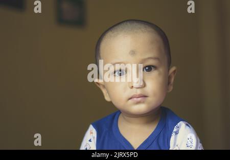 Grazioso bambino indiano calvo in camicia blu e bianca che guarda lontano. Colpo alla testa e alla spalla. Primo piano. Foto Stock