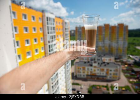 un bicchiere di caffè sulla mano di un uomo si stese nella finestra sullo sfondo di un edificio di appartamenti Foto Stock