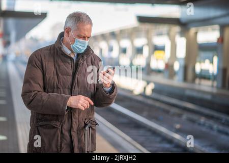 Uomo d'affari mascherato che utilizza il suo smartphone mentre attende il concetto di mobilità in treno, covid e coronavirus Foto Stock