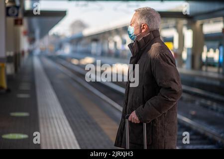 Uomo d'affari senior mascherato in attesa del concetto di mobilità del treno, del covid e del coronavirus Foto Stock