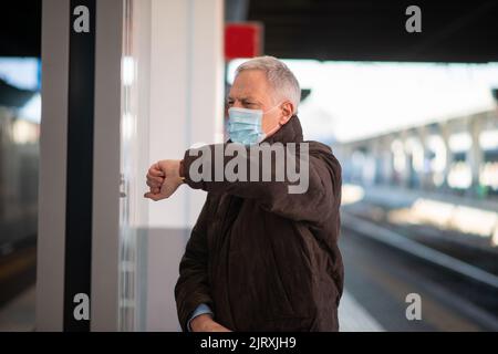 Uomo d'affari senior mascherato in attesa del concetto di mobilità del treno, del covid e del coronavirus Foto Stock
