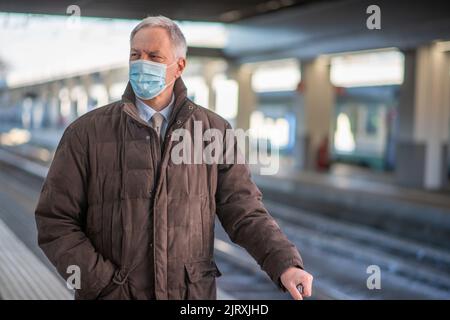 Uomo d'affari senior mascherato in attesa del concetto di mobilità del treno, del covid e del coronavirus Foto Stock