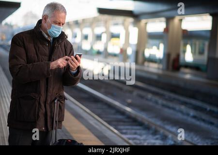 Uomo d'affari mascherato che utilizza il suo smartphone mentre attende il concetto di mobilità in treno, covid e coronavirus Foto Stock