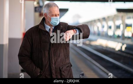 Uomo d'affari senior mascherato in attesa del concetto di mobilità del treno, del covid e del coronavirus Foto Stock