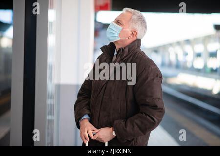 Uomo d'affari senior mascherato in attesa del concetto di mobilità del treno, del covid e del coronavirus Foto Stock